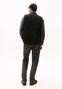 Schwarzes Baumwollwestenoberteil über einem grauen Langarmshirt, kombiniert mit dunklen Jeans und schwarzen Sneakers. Die Rückansicht zeigt ein minimalistisches Design und eine passende Passform.