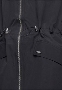 Schwarzer Jackenstoff mit zentralem silbernem Reißverschluss, verstellbaren Kordelzügen mit Stoppern und einer Tasche mit kleinem Metallmarkenetikett.