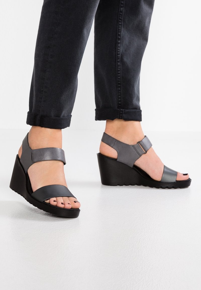 Sandales compensées noires et grises avec une large bride et une bande à la cheville. La semelle présente un motif texturé antidérapant, et le design inclut des orteils ouverts.