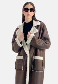 Bruine jas van imitatieleer met een zachte crème-kleurige kraag en zakaccenten. Heeft een open voorkant, grote opgestikte zakken en een losse pasvorm.