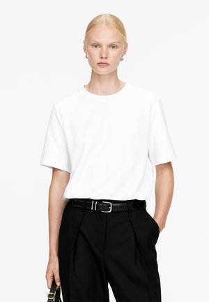 Camiseta blanca de algodón con escote redondo y mangas cortas, combinada con pantalones negros de cintura alta y pliegues, junto con un cinturón negro.