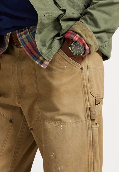 Pantalones de trabajo marrón claro con una tela texturizada, con bolsillos, costuras visibles y manchas de pintura blanca. Se lleva una chaqueta verde y una camisa a cuadros debajo.