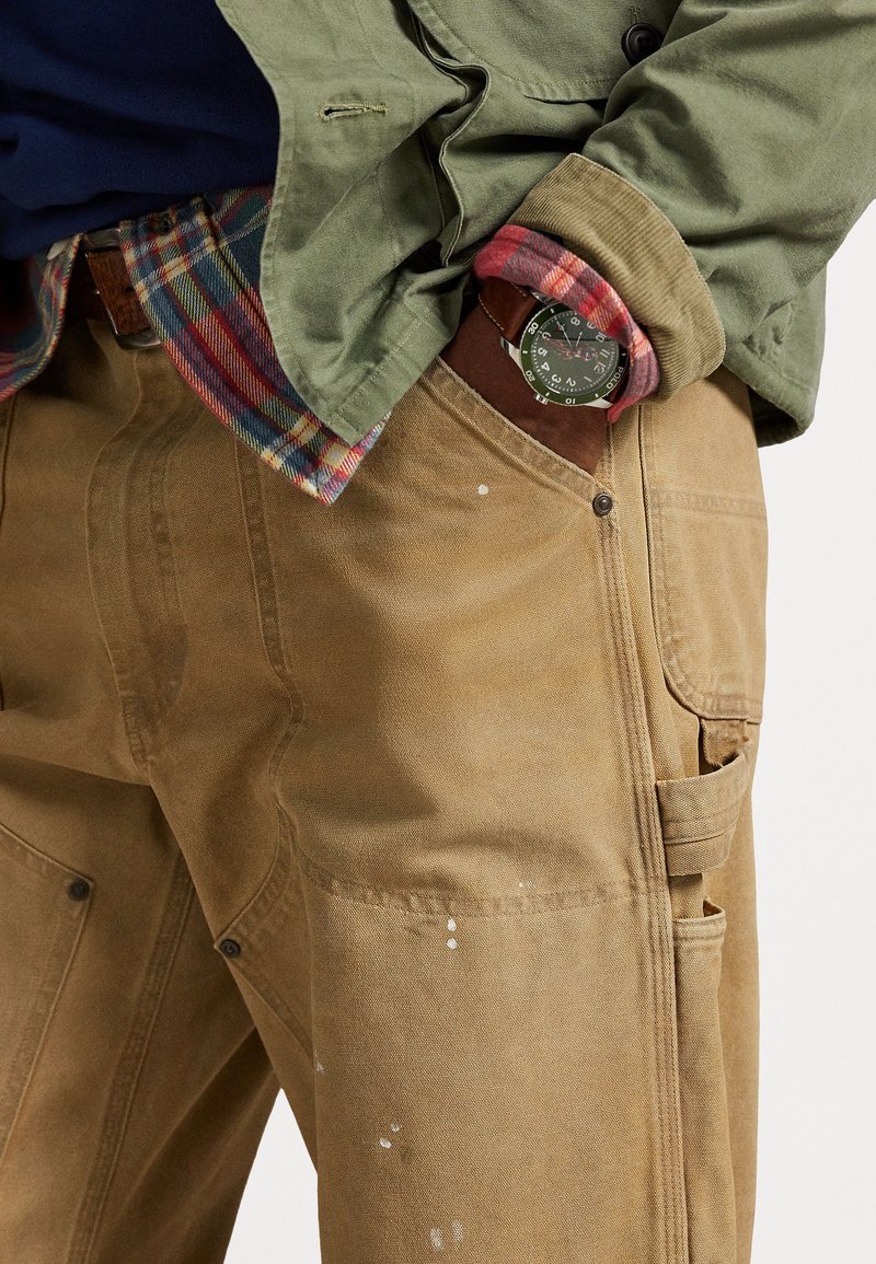 Pantaloni da lavoro marrone chiaro con un tessuto testurizzato, dotati di tasche, cuciture a vista e macchie di vernice bianca. Sotto è indossata una giacca verde e una camicia a quadretti.