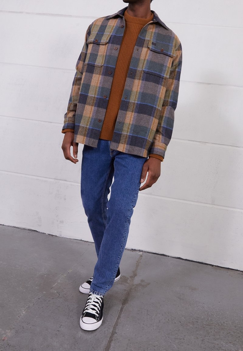 Rutig flanellskjorta i brunt, blått och grönt med två bröstfickor, kombinerad med en brun ribbad tröja och blå jeans. Svarta sneakers.