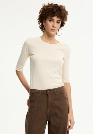 Mujer con pelo rizado que lleva una camiseta crema de manga corta y pantalones marrones de talle alto, de pie frente a un fondo claro y liso.