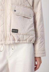 Jachetă umplută de culoare bej deschis, cu un model de tip pătrățel, închidere cu fermoar, buzunar cu capse și tiv ajustabil cu șnur. Logo-ul Barbour International este vizibil.