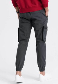 Graue Cargo-Hosen mit elastischen Bündchen, ausgestattet mit seitlichen Taschen und einer Reißverschlusstasche. Hergestellt aus strapazierfähigem Stoff, kombiniert mit weißen Sneakern.