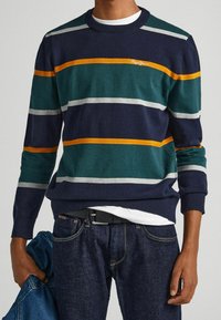 Man i marint, grönt, vitt och orange randig långärmad tröja, mörka jeans med svart bälte, hållande en blå denimjacka i vänster hand.