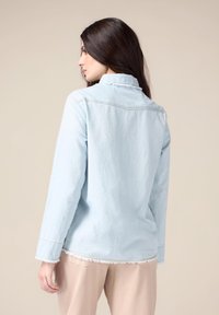 Light blue denim shirt with long sleeves, frayed hem, and subtle stitching details. The fabric has a soft, textured appearance.