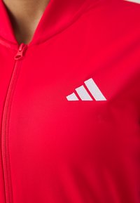 Červená športová bunda so zipsom na prednej strane, vyrobená z hladkého materiálu. Na ľavej strane sa nachádza biele logo Adidas.