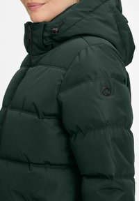 Frau in einer dunkelgrünen gepolsterten Jacke mit hohem Kragen und Kapuze, die ein schwarzes rundes Logo auf dem oberen Ärmel zeigt.