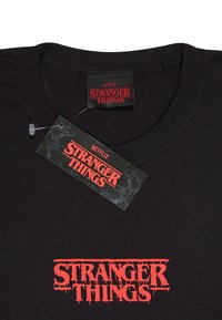 Černé bavlněné tričko s červeně potisknutým logem "STRANGER THINGS" s efektem kapání a štítkem se podobnými designovými prvky.