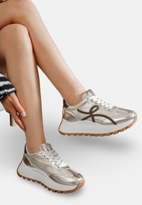 Baskets argentées métalliques avec une tige en maille texturée, des lacets bruns contrastants et une semelle plate blanche avec une semelle en caoutchouc brun.