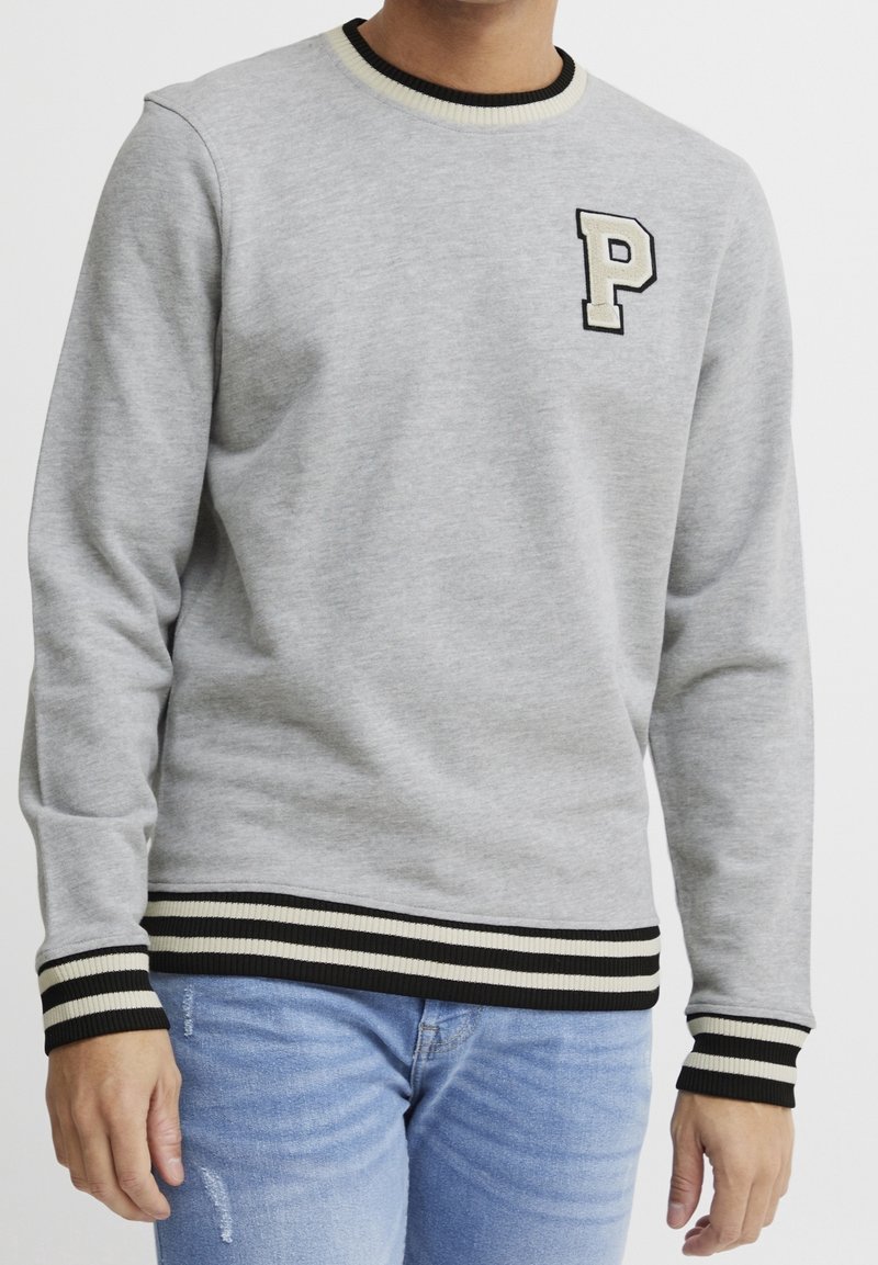 Homme portant un sweat-shirt gris clair avec des manches et un bas à rayures noires et blanches, et un patch blanc avec la lettre "P" sur la poitrine.