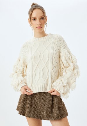 Cremfarbener Strickpullover mit strukturierten Zopfmuster und fransigen Ärmeln, kombiniert mit einem braunen Hahnentrittmuster-Rock in Glockenform.