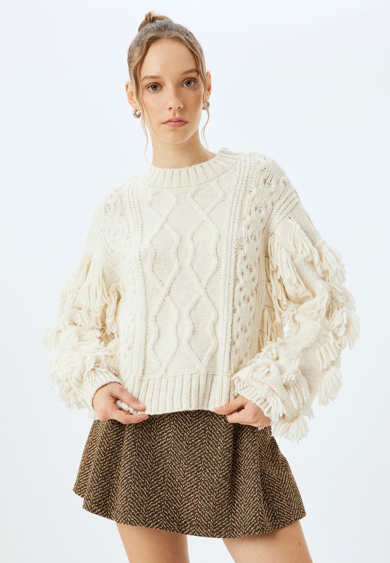 Kremowy sweter dzianinowy z teksturowanymi wzorami w warkocze i frędzlami na rękawach, połączony z brązową spódnicą w kratkę typu houndstooth o rozszerzanym kroju.