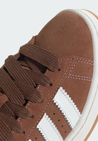 Brown suede sneaker featuring white leather stripes, textured laces, and a gum rubber sole. Notable stitching detail along the upper.