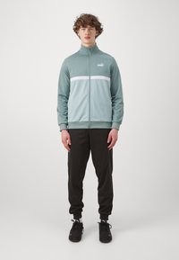 Lys grøn zip-up træningsjakke med hvid vandret stribe, parret med sorte tætsiddende bukser og sorte atletiske sko, stående på en ensfarvet baggrund.