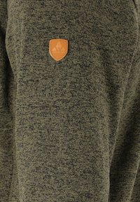 Mörkgrön, strukturerad tyg har en läderpatch med en sköldlogotyp. Designen inkluderar subtila variationer i färg och mönster.