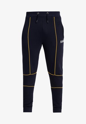Pantaloni da jogging blu navy con dettaglio piping giallo, vita con coulisse, polsini a coste e testo bianco parziale sulla parte superiore della coscia.