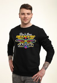 Svart sweatshirt med en färgglad grafisk tryck som har texten "Don't Stop BELIEVIN'" samt en design med en fågel, en planet och vingar.
