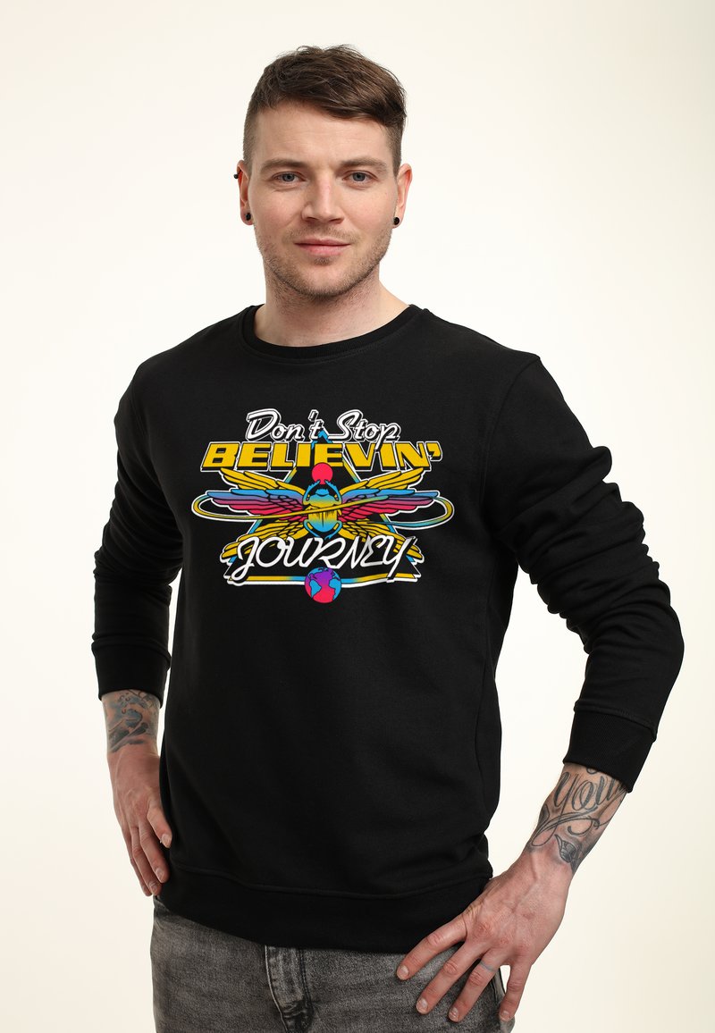 Svart sweatshirt med en färgglad grafisk tryck som har texten "Don't Stop BELIEVIN'" samt en design med en fågel, en planet och vingar.