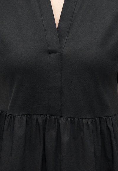 Μαύρο φόρεμα με V-λαιμόκοψη και λεπτομέρεια σούρωσης στη μέση, που δείχνει το πάνω μέρος του κορμού και την υφή του υφάσματος.