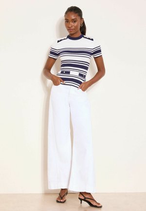 Kobieta stoi przy białej ścianie, ubrana w granatowo-biały pasiasty krótki rękaw, białe spodnie o szerokich nogawkach oraz czarne sandały na obcasie.