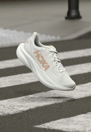 Witte Hoka hardloopschoen met beige logo, zwevend boven een voetgangersoversteekplaats op asfalt.
