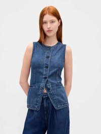 Young woman with straight red hair wearing sleeveless buttoned denim vest and loose dark denim pants on plain background