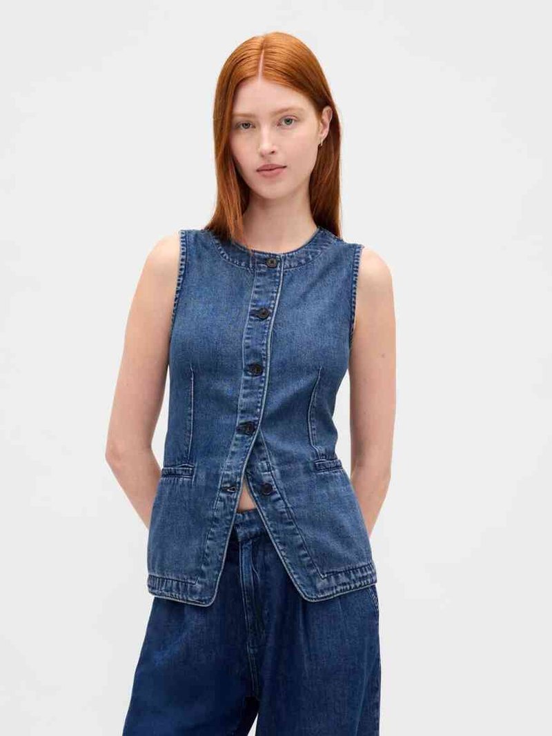 Young woman with straight red hair wearing sleeveless buttoned denim vest and loose dark denim pants on plain background