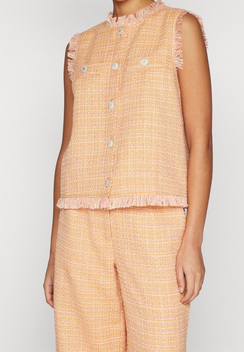Bezramenná tweedová vesta v broskvové barvě s texturovaným vzorem, stříbrnými knoflíky a třepícími se okraji na límci a spodním lemu. Viditelné kalhoty ve stejném stylu.
