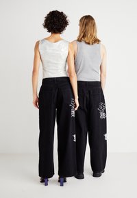 Jean noir à jambes larges avec broderie blanche sur les poches, associés à un haut en sequins argentés et un débardeur gris. Les deux portent des chaussures à talons carrés.