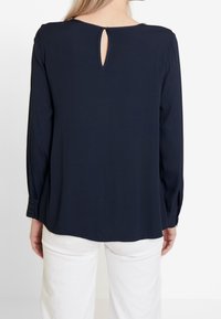 Frau von hinten gesehen, die eine dunkelblaue Bluse mit einem Ausschnitt am Hals trägt, kombiniert mit weißen Hosen. Glattes Material, lange Ärmel.