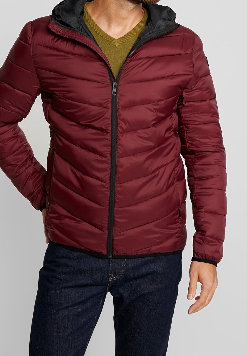 Burgundská péřová bunda s vypasovaným střihem a černou kapucí. Texturovaný, prošívaný povrch s předním zipem a bočními kapsami.