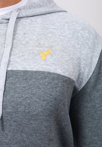Grå og lys grå hettegenser med en myk tekstur, med en brodert gul fuglelogo på brystet og en snor til hetten.