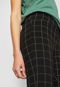 Svarta leggings med ett vitt rutmönster, tillverkade av stretchigt material. Närbild av sidodetaljen med synlig vit sömnad.