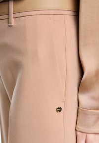 Hellrosa maßgeschneiderte Hose mit glatter Textur, verziert mit einem goldfarbenen Metallakzent und einem eleganten Hosenbund, detaillierte Steppnähte entlang der Seite.