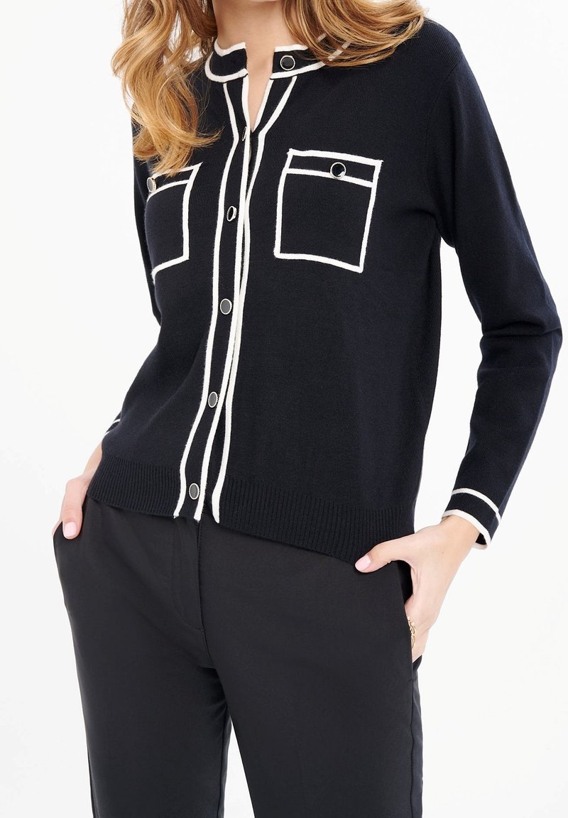 Cardigan en tricot marine avec passepoil blanc, deux poches poitrine et patte de boutonnage ; design classique avec une finition texturée.