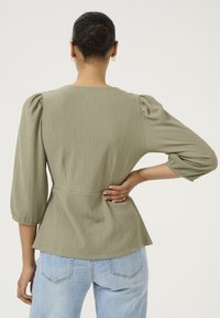 Top peplum verde oliva con tessuto strutturato, maniche a sbuffo e vita aderente. Appare con una vestibilità rilassata, abbinato a jeans azzurri.