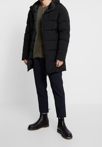 Sort puffermantel med lynlås, hætte og quiltet design. Båret over en grøn tekstureret top og parret med marineblå bukser og sorte støvler.
