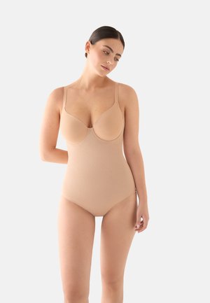 Mujer de cabello oscuro recogido en un moño elegante, lleva un body ceñido de color nude con copas con aro, de pie contra un fondo claro y sencillo.