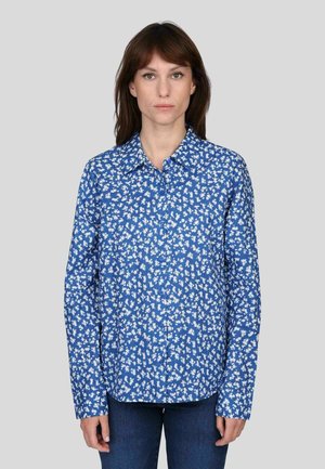 Chemise bleu à boutons avec un motif floral en blanc. Dotée d'un col évasé et de manches longues. Portée avec un jean bleu, présentée sur un fond gris.