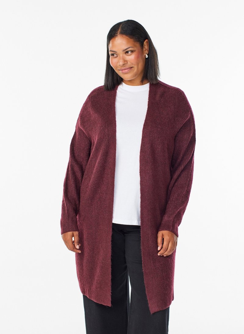 Donna che indossa un cardigan lungo color amaranto con apertura frontale, camicia bianca e pantaloni neri, in piedi su sfondo bianco.