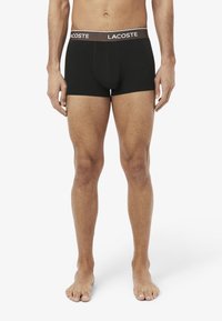 Mannelijke torso en benen met zwarte Lacoste boxershorts en een bruine tailleband met het Lacoste-logo, staand op blote voeten tegen een witte achtergrond.