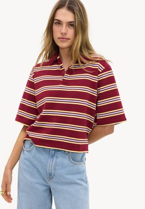 Mujer joven con una camiseta tipo polo de rayas burdeos con líneas amarillas, blancas y azul claro, combinada con vaqueros de talle alto azul claro.