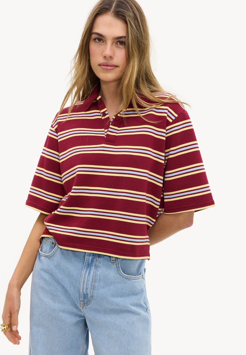 Jeune femme portant un polo rayé bordeaux avec des lignes jaunes, blanches et bleu clair, associé à un jean taille haute bleu clair.