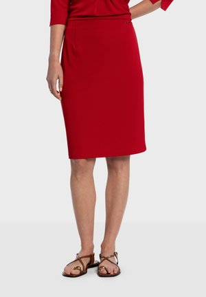 Červená ceruzková sukňa do kolien vyrobená z hladkej látky, s jemným detailom vrecka a bočným zapínaním. Nosená s hnedými sandálmi.