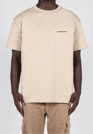 Mand iført beige T-shirt med korte ærmer med lille sort "MJGONZALES" tekst på brystet og lysebrune cargobukser.
