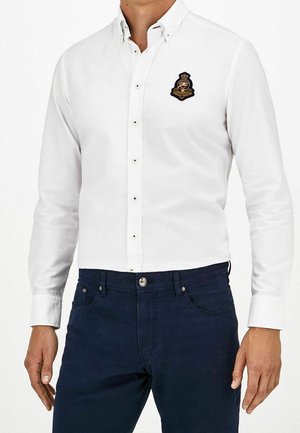 Mężczyzna w białej koszuli na guziki z naszywką herbu na piersi i ciemnoniebieskich dżinsach, stojący z rozluźnionymi ramionami.